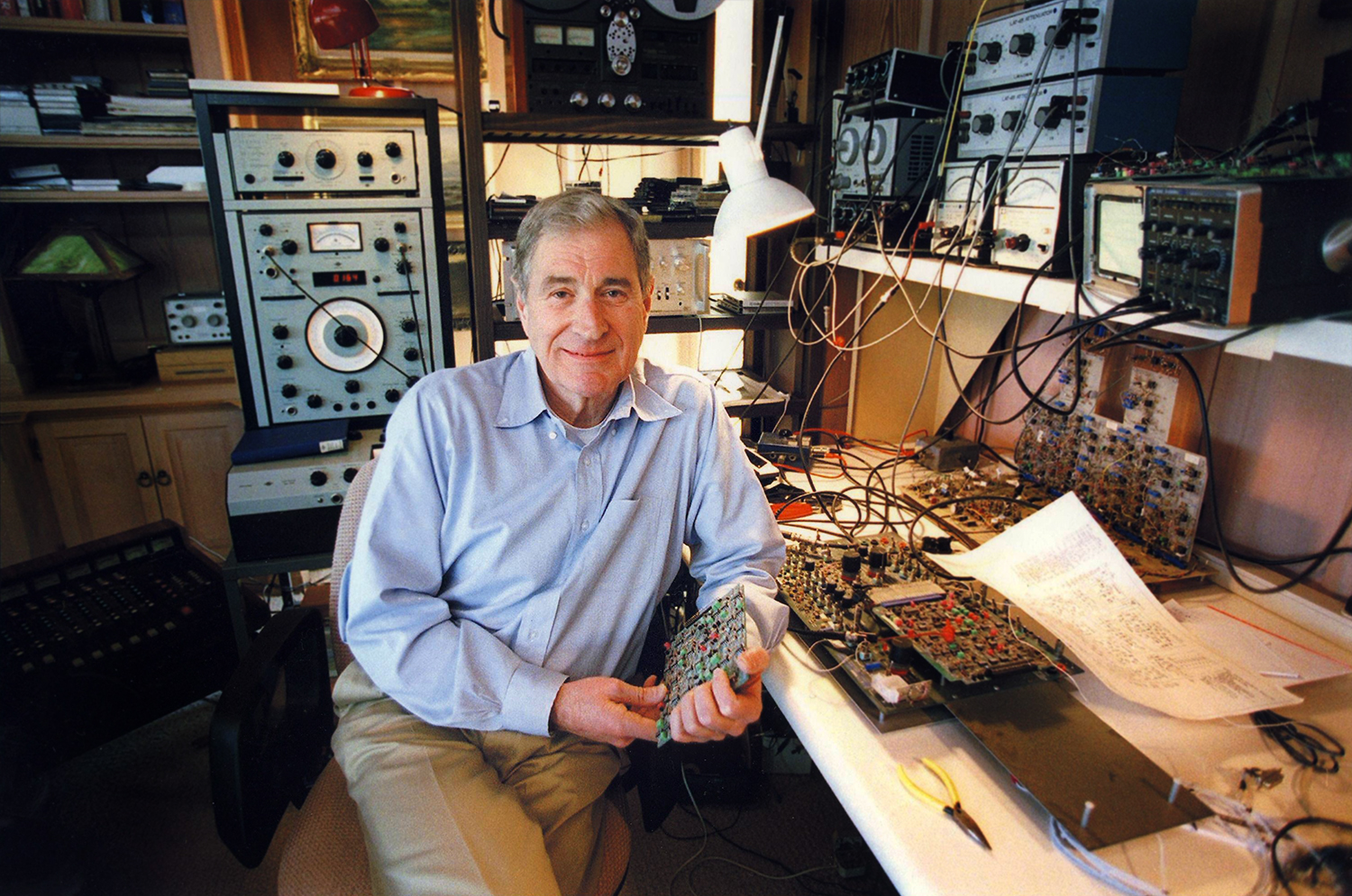 Ray Dolby seated in his workshop holding one of his circuit boards.